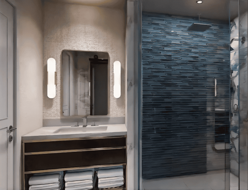 Modern bathroom with a double sink vanity, mirror with two wall lights, stacked towels below, and a glass-enclosed shower with blue tiled walls and a ceiling-mounted showerhead.