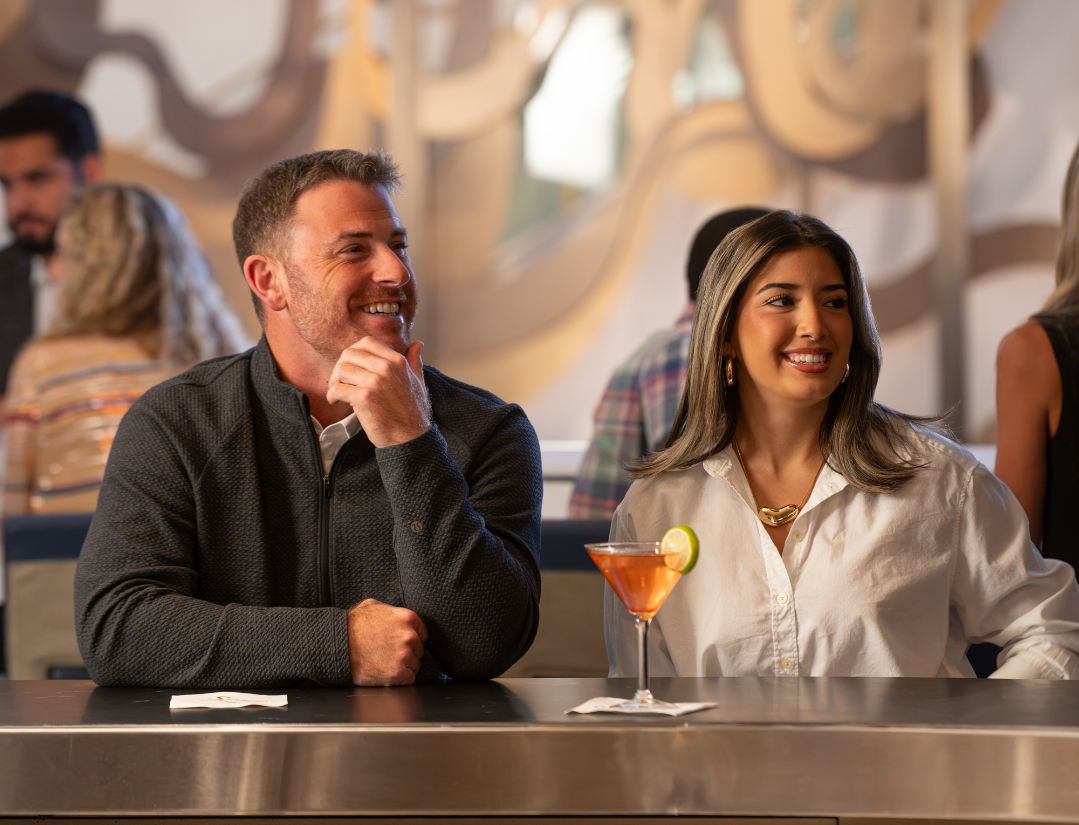 A man and a woman sit at a bar, smiling and looking to the side. A cocktail with a lime garnish is in front of the woman. Other people are in the background.