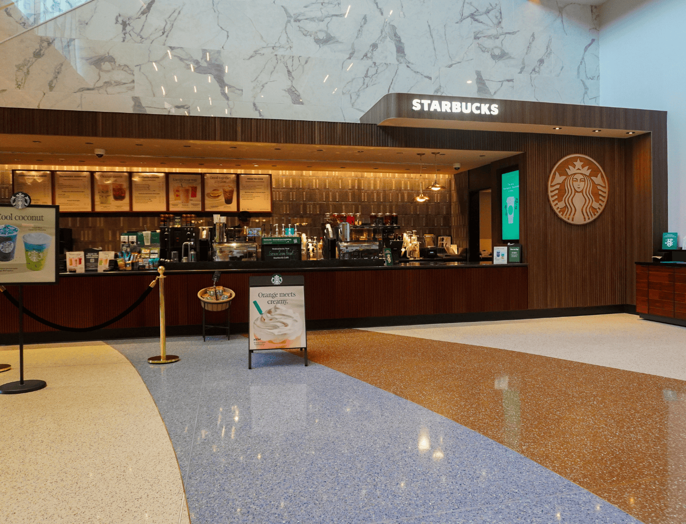 Starbucks coffee shop counter with menu boards, promotional signs, and coffee machines, set in a modern space with marble walls and terrazzo flooring.