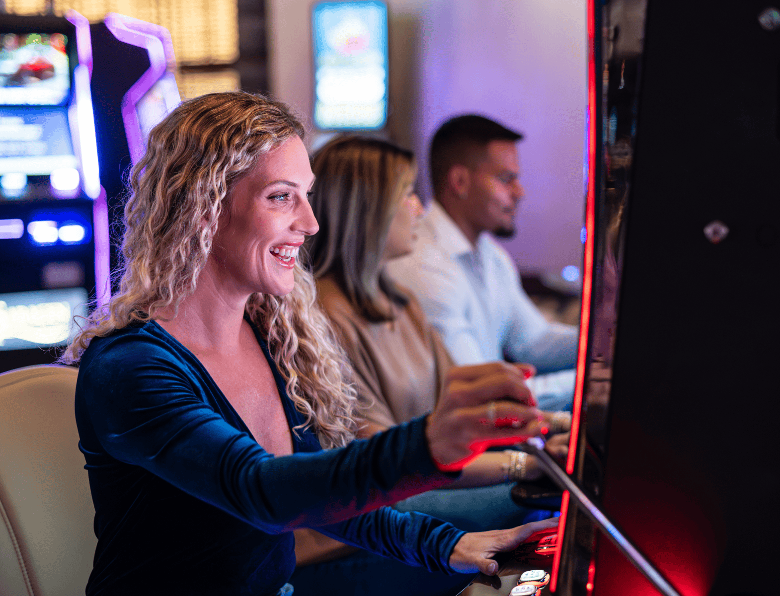 A woman in a blue top plays a slot machine at a casino, smiling. Two other people play machines beside her. Bright gaming screens are visible in the background.