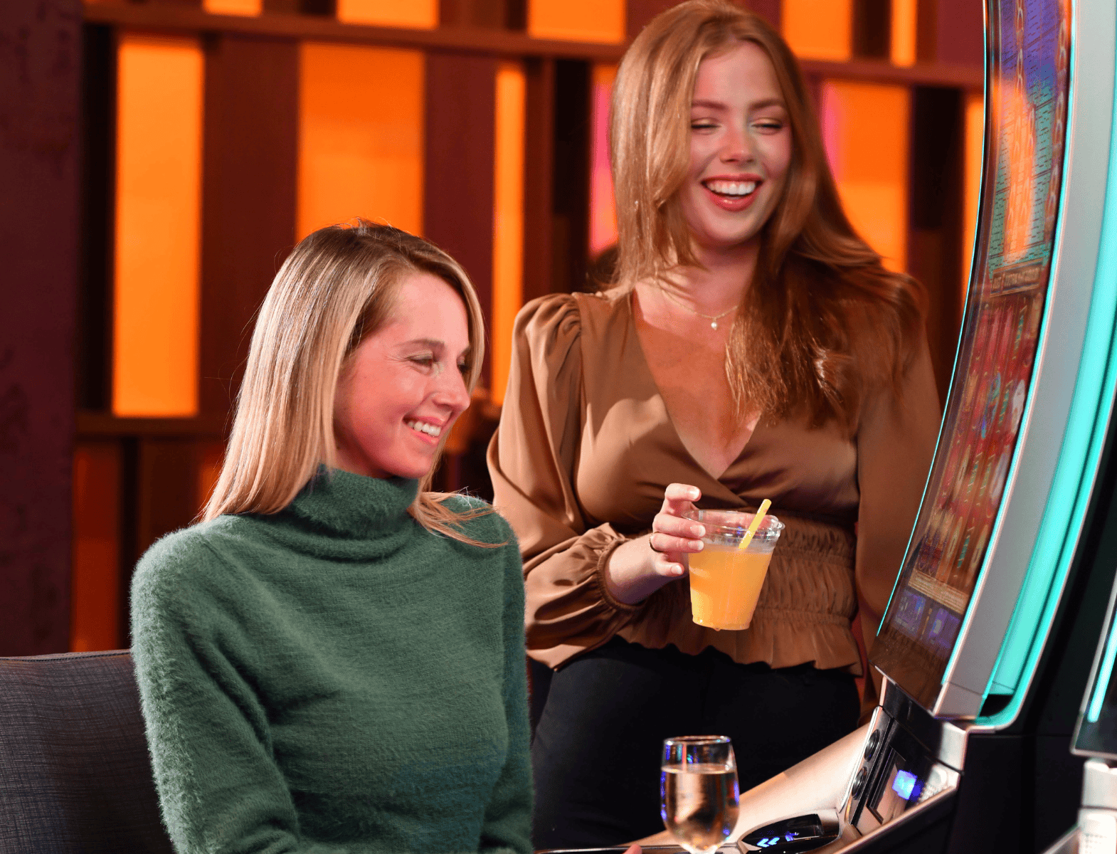 Two women at a slot machine, one seated and smiling, the other standing with a drink. There is a glass of white wine on the counter in front of them.