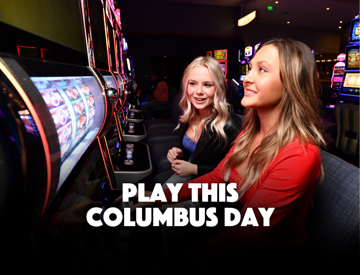 Two women sit at slot machines in a casino, smiling, with the text “Play This Columbus Day” displayed at the bottom of the image.
