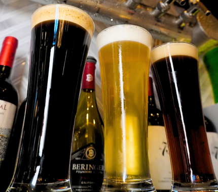 Three tall glasses of beer—dark, light, and amber—stand in front of several wine bottles on a bar counter.