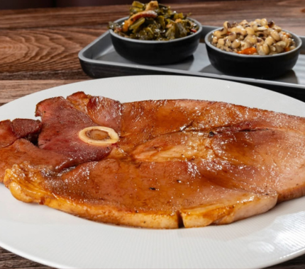 A glazed ham steak on a white plate, served with side bowls of greens and black-eyed peas.