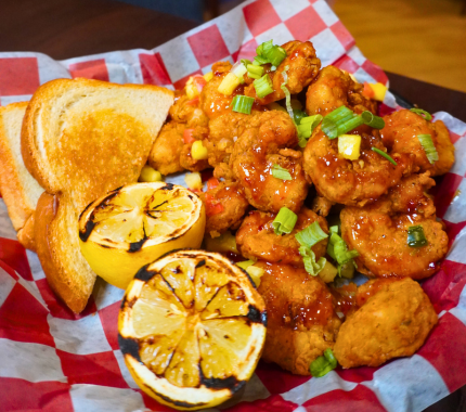 A basket of fried shrimp drizzled with sauce and topped with green onions, served with two slices of toasted bread and two charred lemon halves.